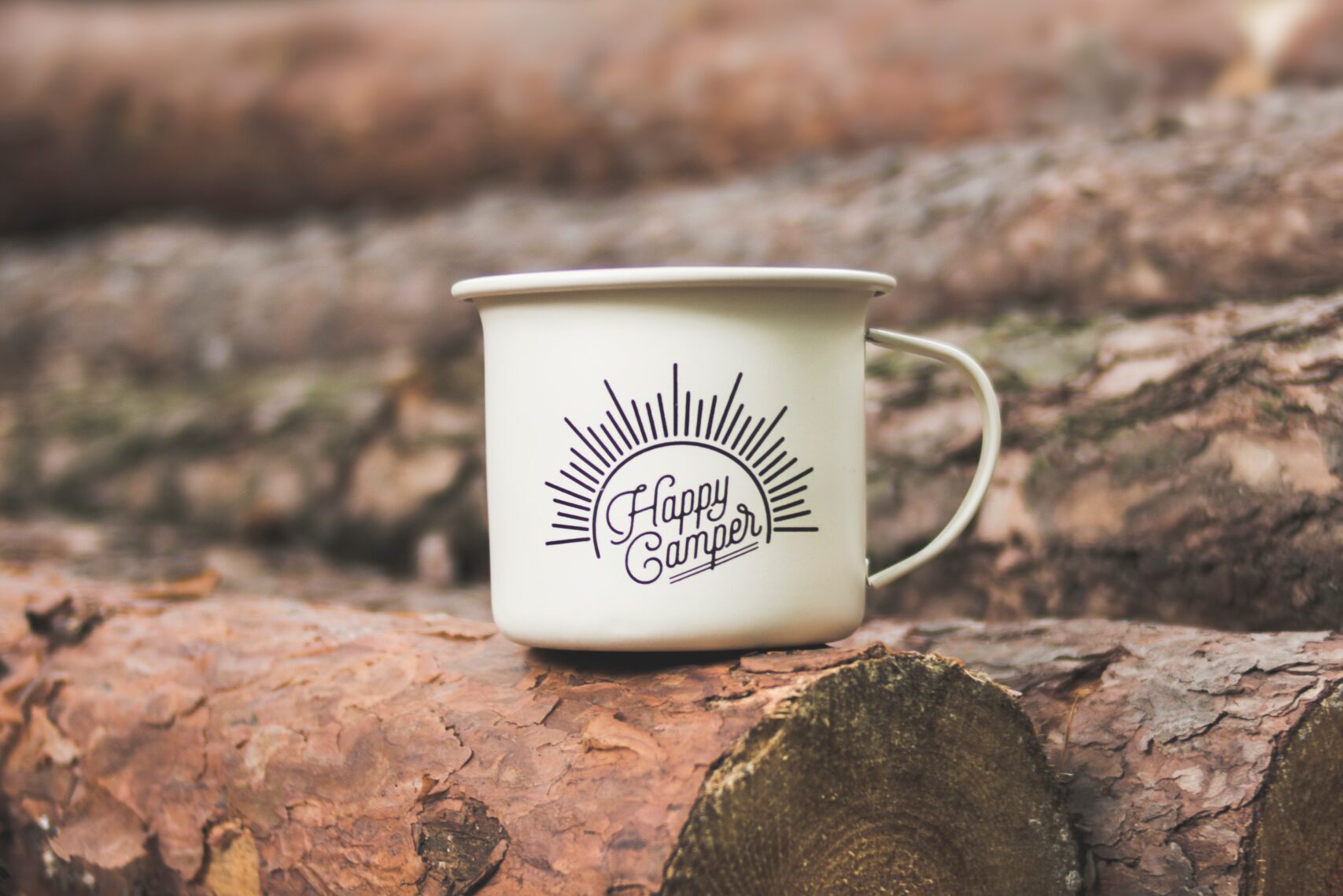 White tin cup sitting on alog in the woods with the words "Happy Camper" printed in black text on it